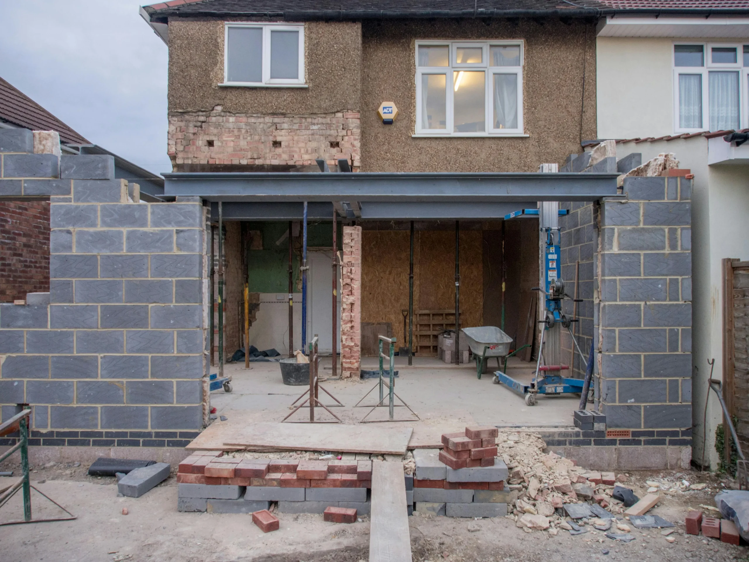 A house under renovation, showing exposed brickwork and construction tools. New walls are partially built with gray blocks. The scene conveys progress.