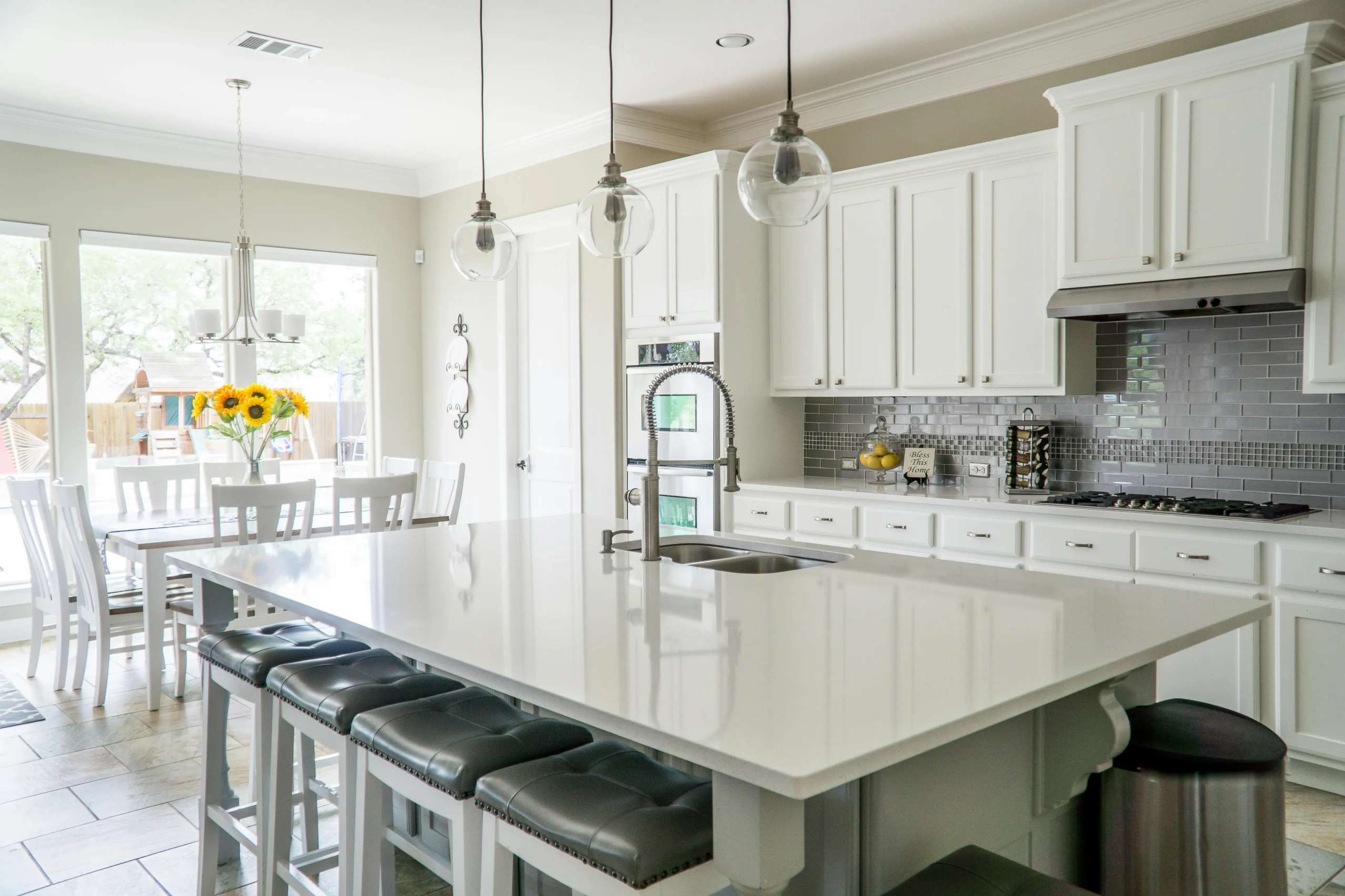 Bright kitchen with white cabinets and island, gray backsplash, and pendant lights. Dining table with sunflowers by large window. Modern and inviting.