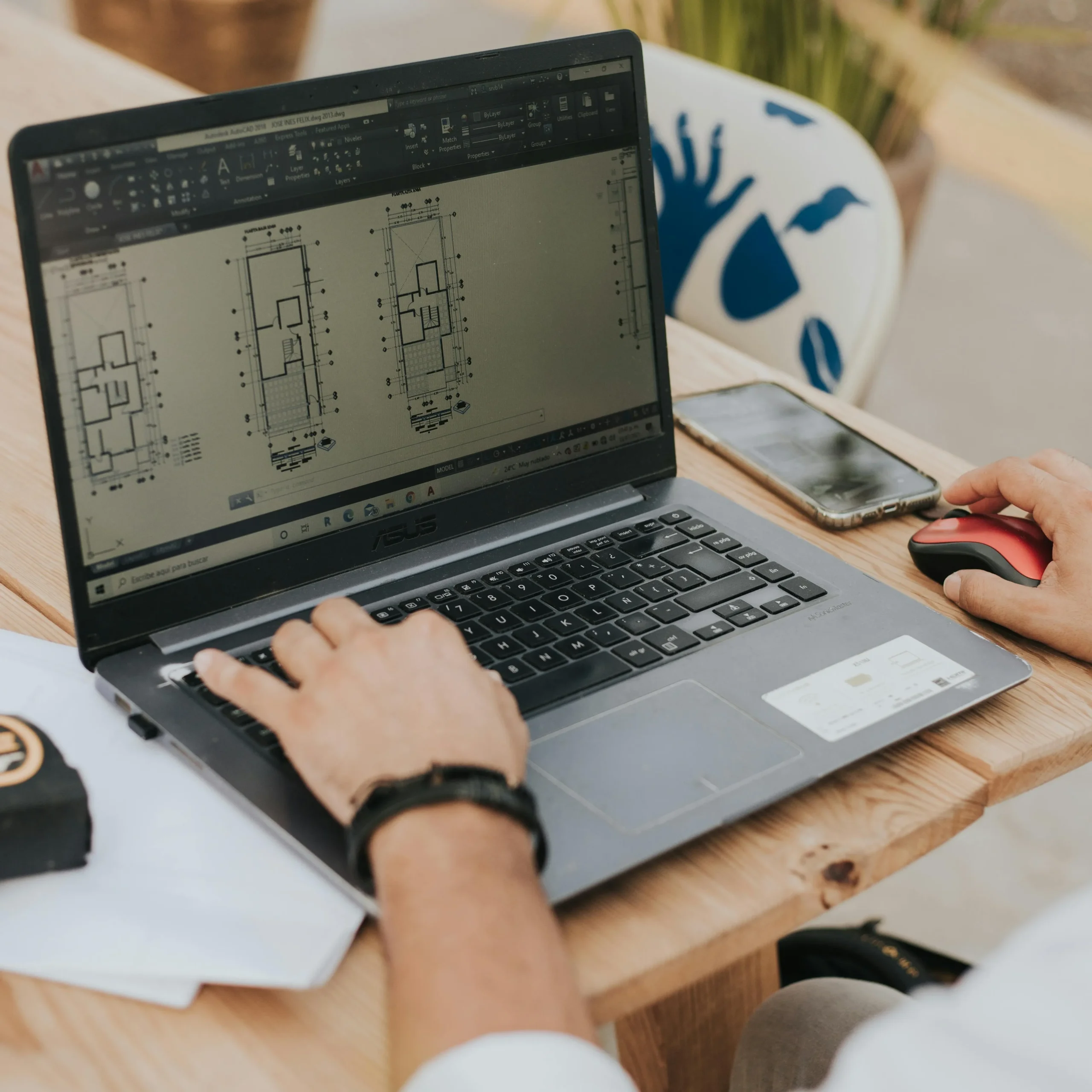 Close-up of a person using a laptop on a wooden table, displaying architectural plans. A smartphone and mouse are nearby, suggesting a focused work setting.