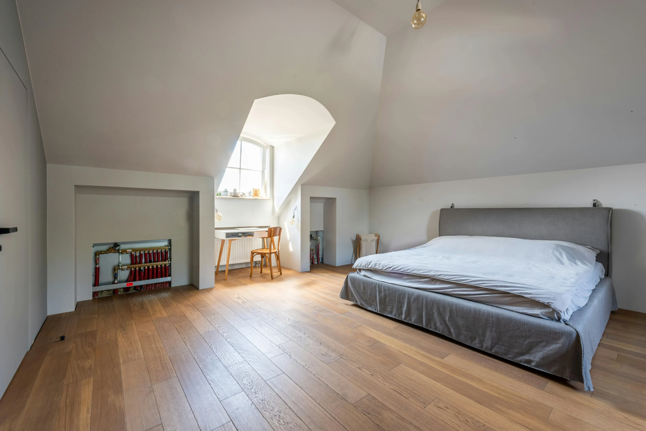 A spacious bedroom with sloped ceiling and wooden floor features a large gray bed on the right. A desk and chair sit under a window on the left, creating a cozy, serene atmosphere.