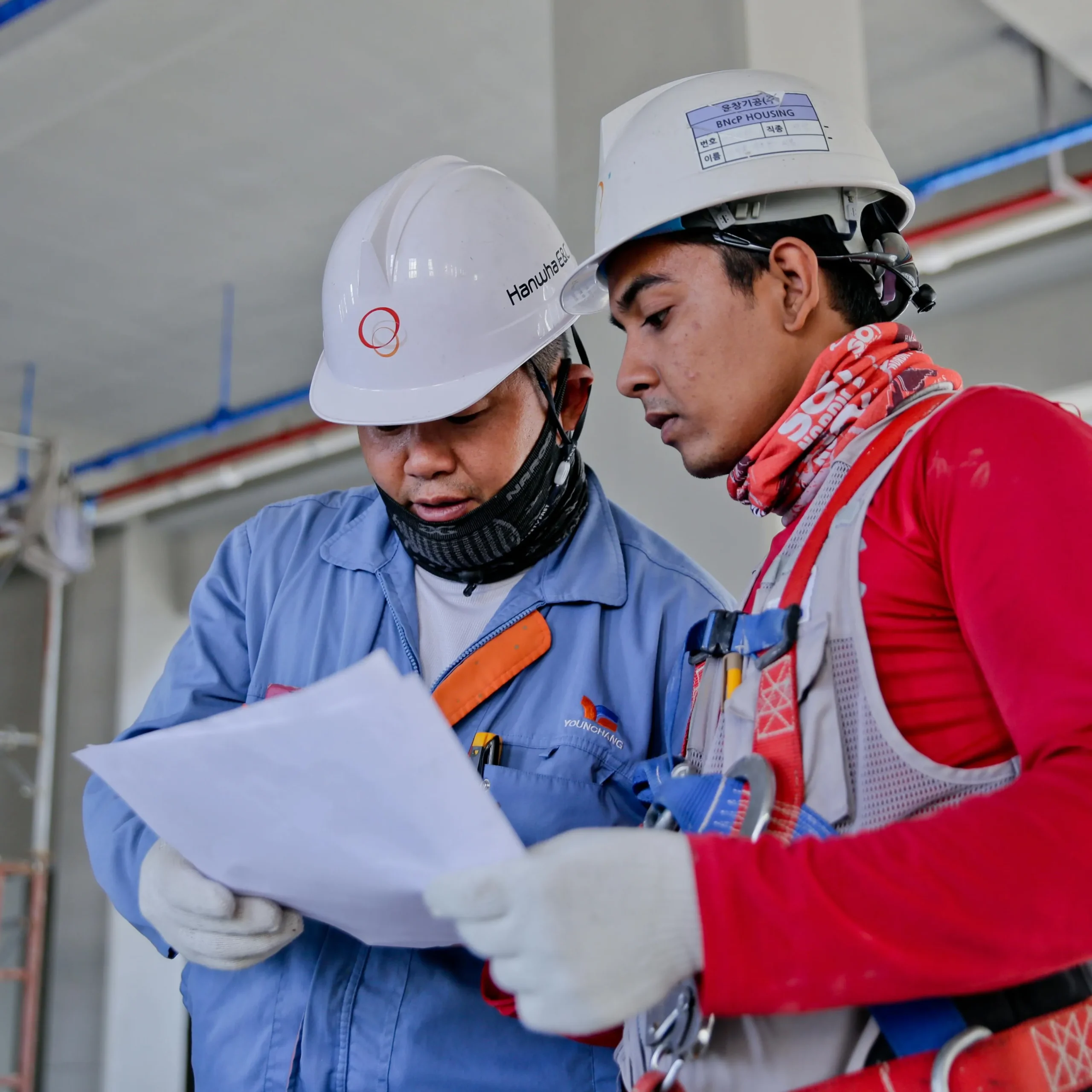 Two construction workers in safety gear and helmets review documents on a building site. They appear focused and engaged, suggesting teamwork and collaboration.