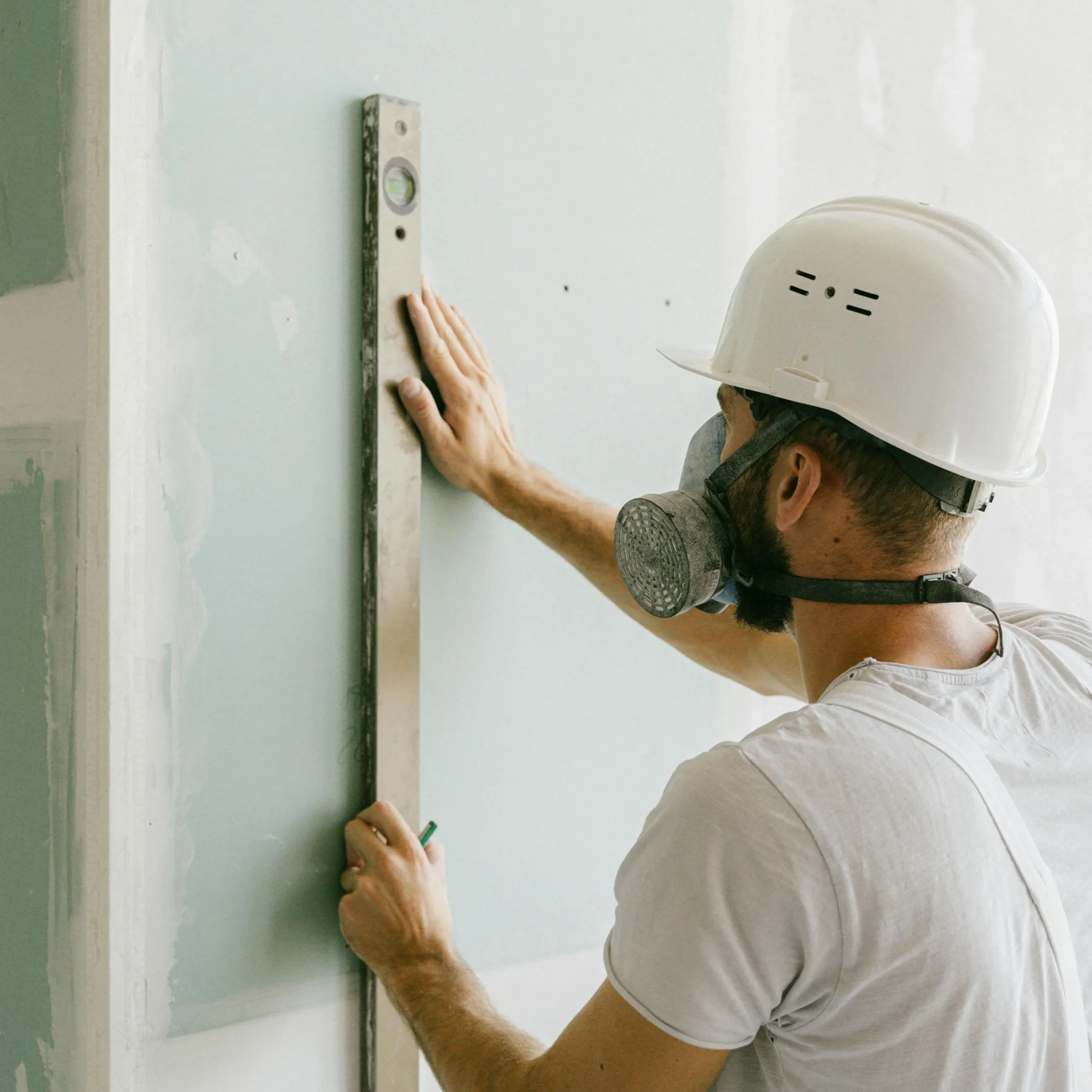 A construction worker in a hard hat and dust mask measures a wall with a metal level. The setting is industrial and focused on precision and safety.
