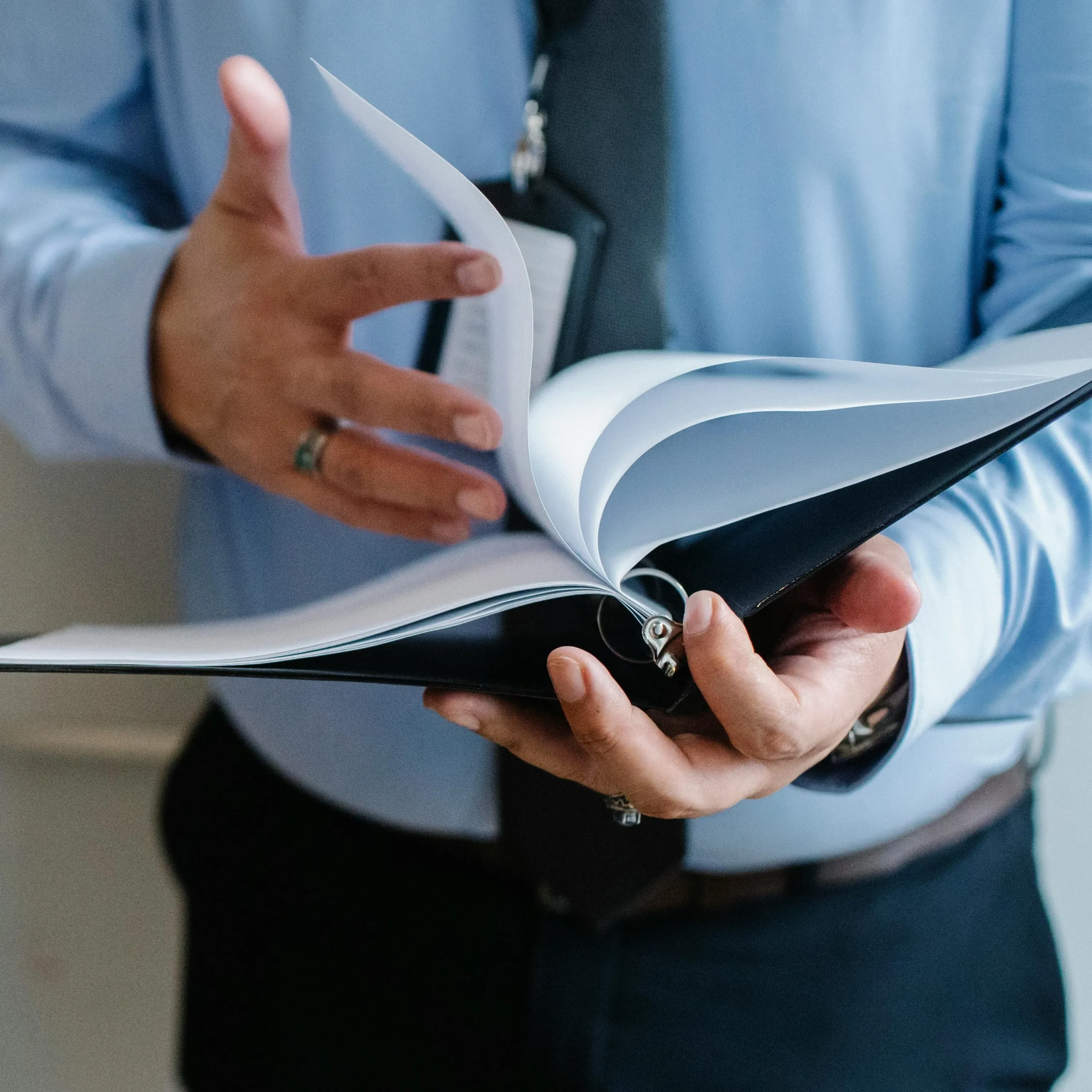 A person in a blue shirt flips through a binder, holding it open with one hand, suggesting focus and engagement. The atmosphere is professional.
