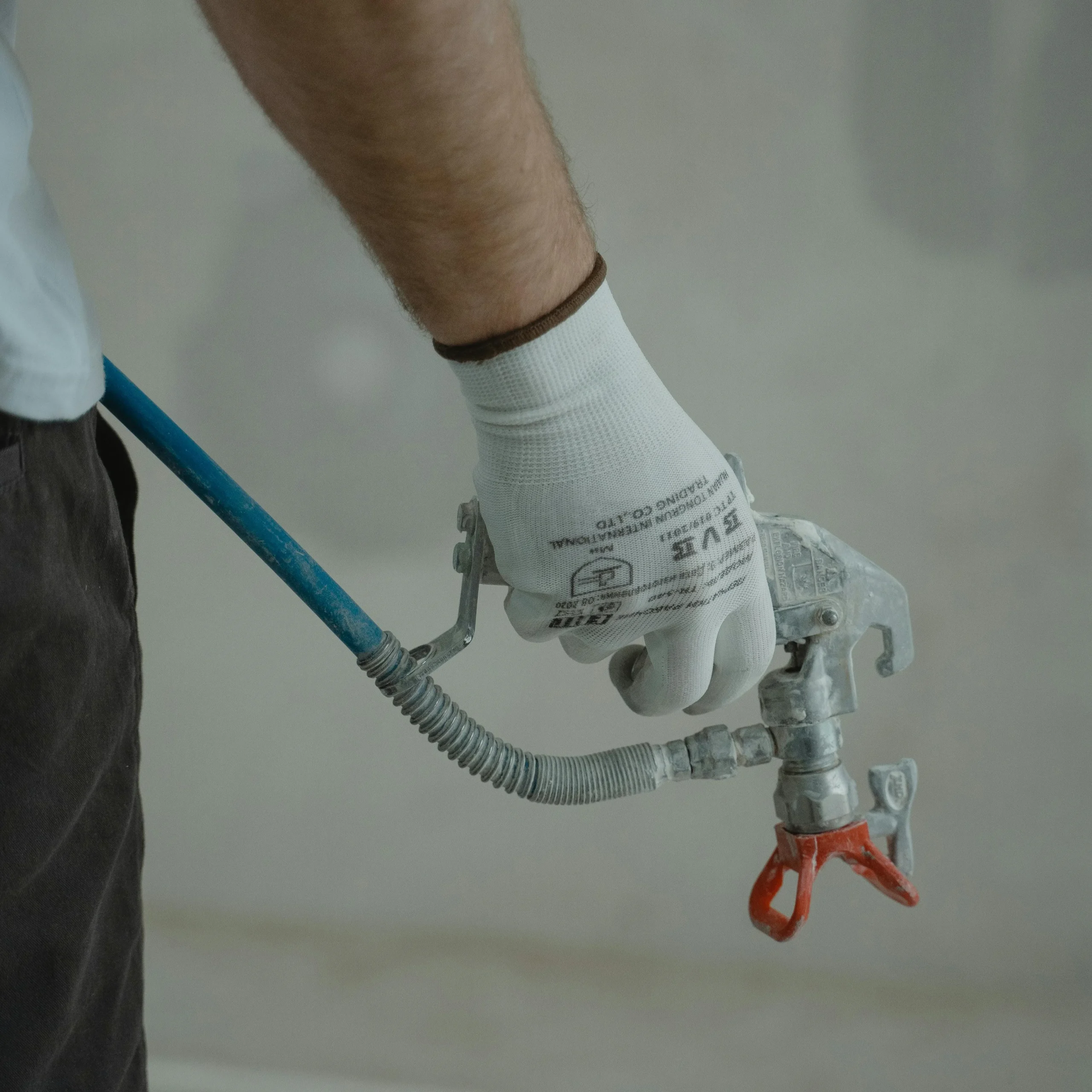 A person wearing white gloves holds a paint spray gun connected to a blue hose, poised to paint. The scene conveys focus and precision in a work setting.