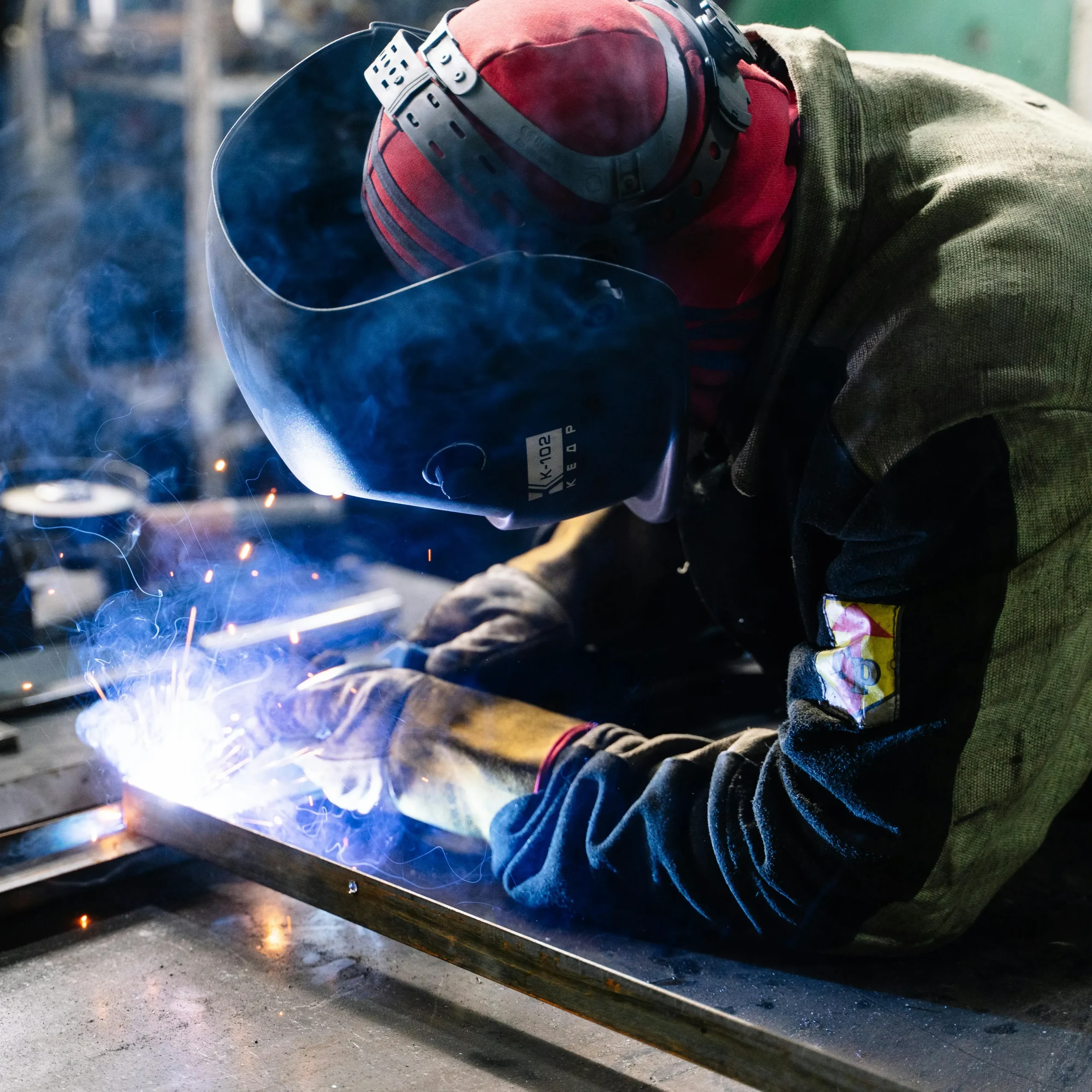 Welder wearing protective gear and mask, focused on welding metal. Sparks illuminate the work area, creating an intense, industrious atmosphere.