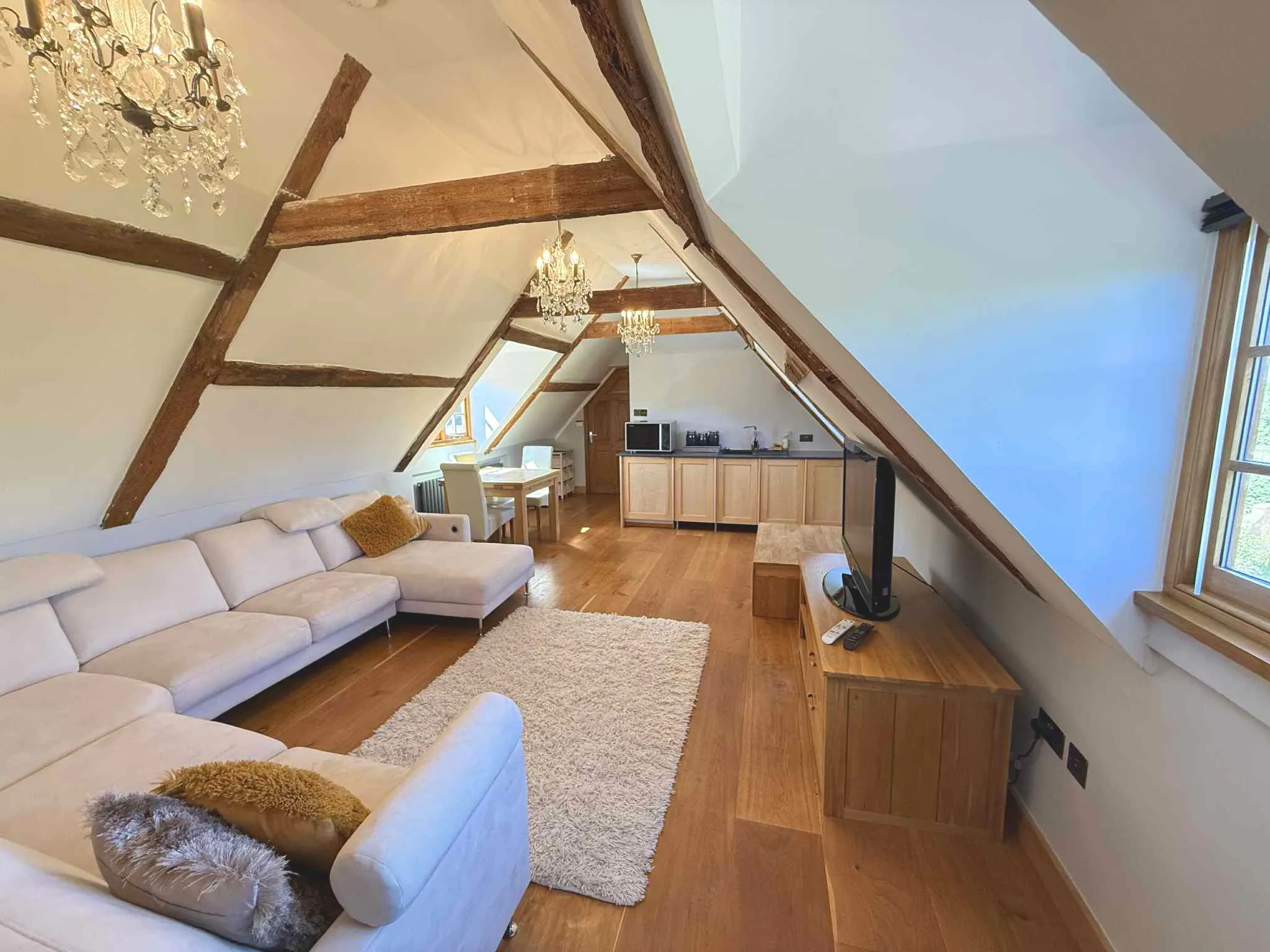 A spacious living area created by a rear dormer loft conversion, featuring exposed wooden ceiling beams, a large corner sofa, oak flooring, and a compact kitchenette setup.